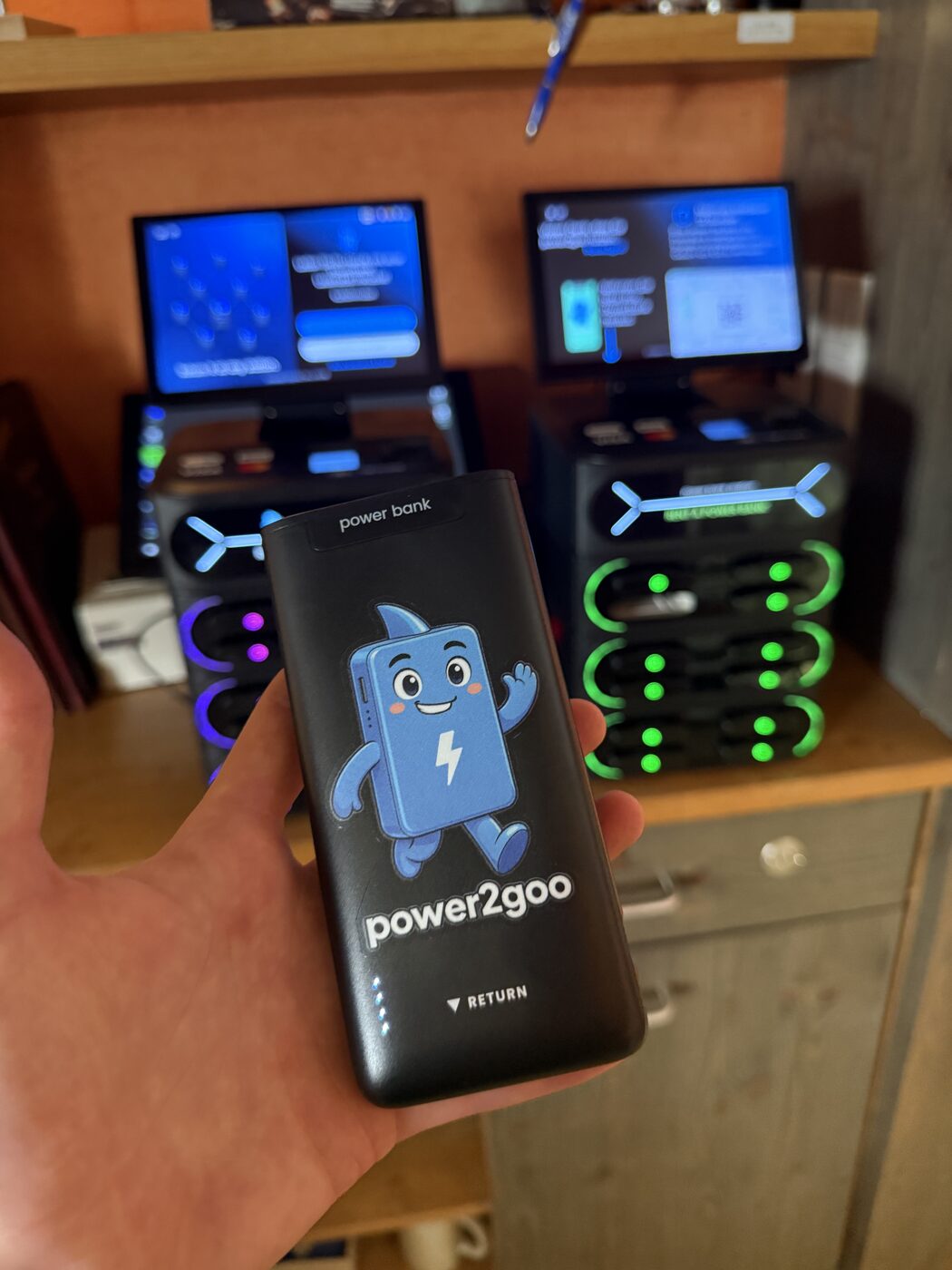 power2goo Powerbank in der Hand, im Hintergrund zwei Stationen